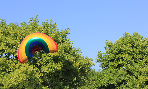 A balloon shaped like a rainbow held up in front of green treetops and a blue sky.