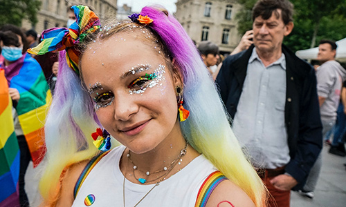 A smiling person in glittery make-up at an outdoor event. They are wearing rainbow earrings, suspenders and hair accessories.