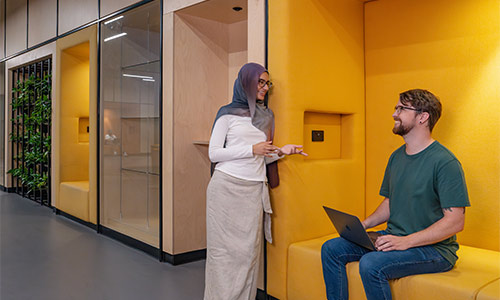 Two students, one standing and one sitting, smile and talk in a yellow seating nook.