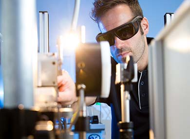 A person wearing a white surgical-style cap and goggles, looking at themselves in the reflection of some equipment.