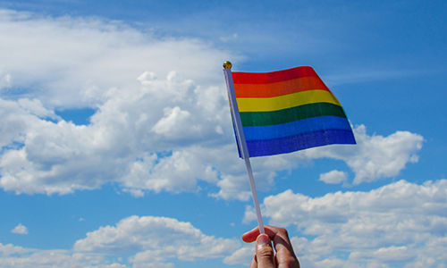 A hand holding a small rainbow pride flag up against a blue sky with white clouds.