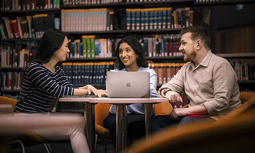 Three students sit around a table in front of tall bookshelves.