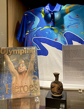 Museum display of a magazine cover showing athlete Grant Hacket in front of a Sydney 2000 Olympic Games volunteer's t-shirt. To the right is a small, ancient Greek oil jug featuring an image of the goddess Nike