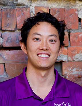 John Lui wearing a purple t-shirt, standing against a brick wall