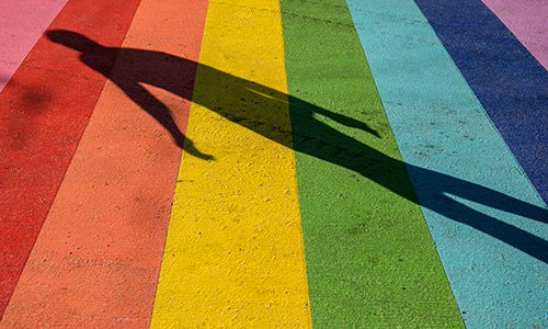 A walkway painted with stripes in the eight colours of the original Pride flag, with a shadow of a person crossing it.