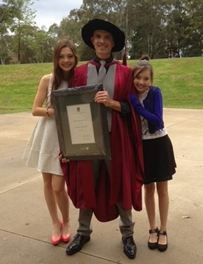 Josh Kidd smiling for a photo, holding an award with his two duaghter on either side of him